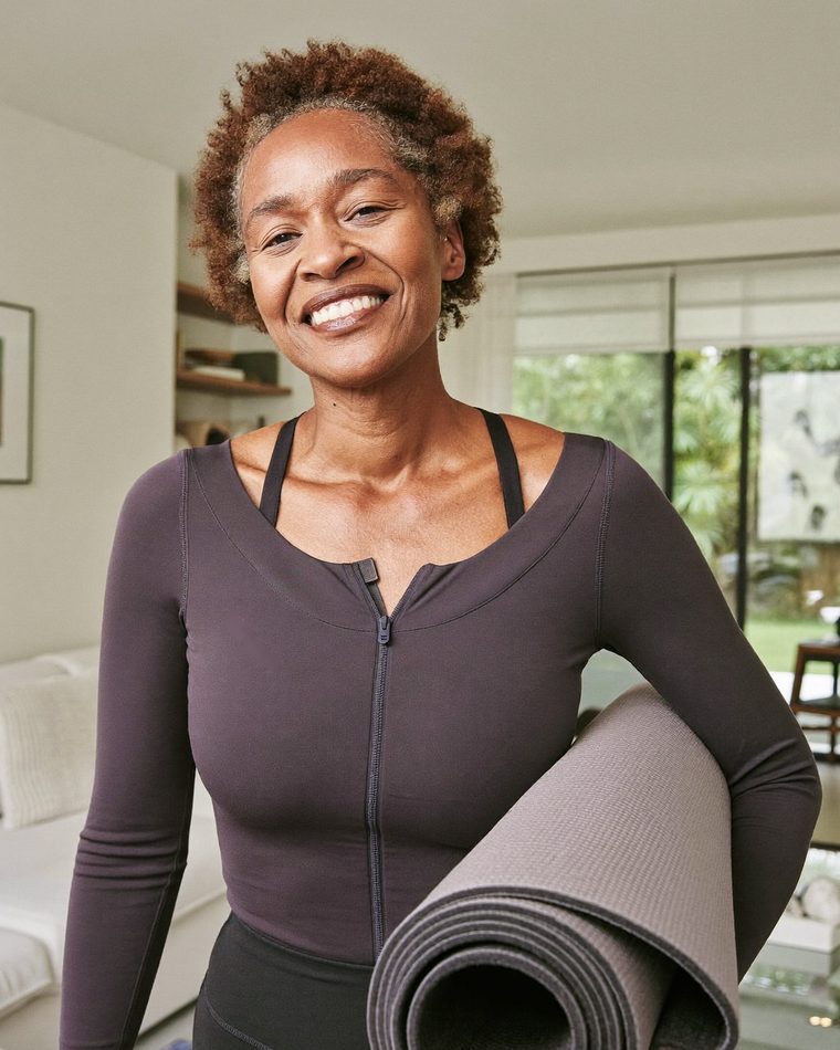 Mulher de cerca de 50 anos sorrindo, em roupa de ginástica, com um tapete de exercícios debaixo do braço, na sala de casa